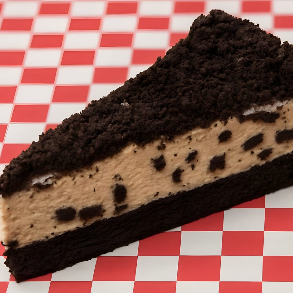 A slice of chocolate cookie cheesecake with a crumbly topping on a red and white checkered surface.